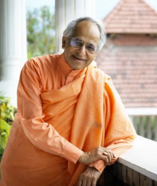 Swami Veda Bharati in posa appoggiato col gomito sul davanzale di una finestra con sullo sfondo un tetto ti un templio